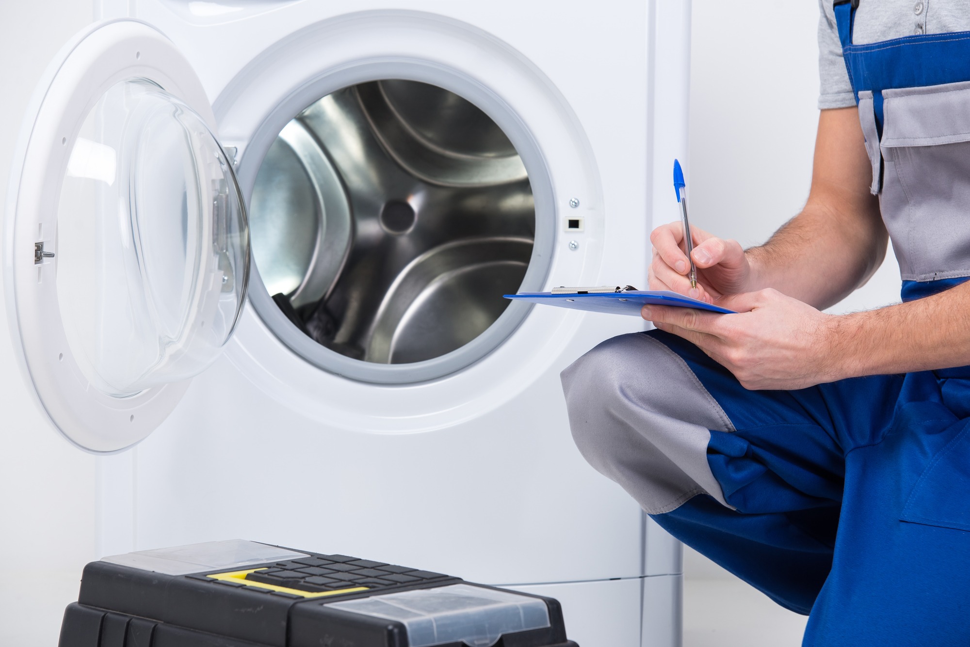 A man in overalls writes on a clipboard beside a washing machine